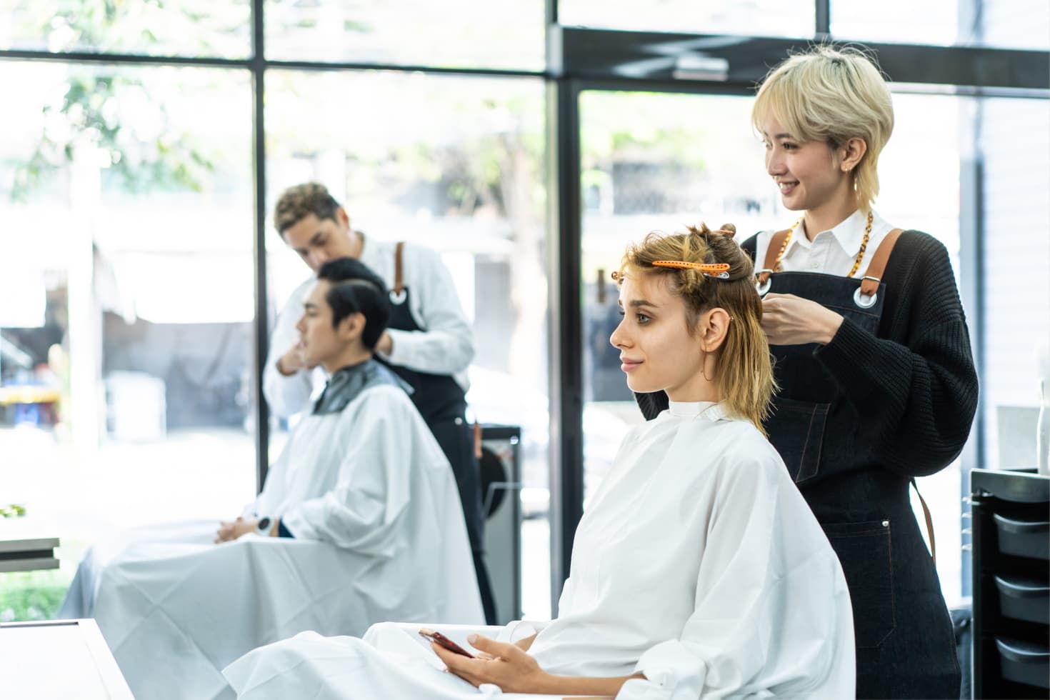 Two stylists cut hair in a well-lit salon, one in the foreground and one in the background.