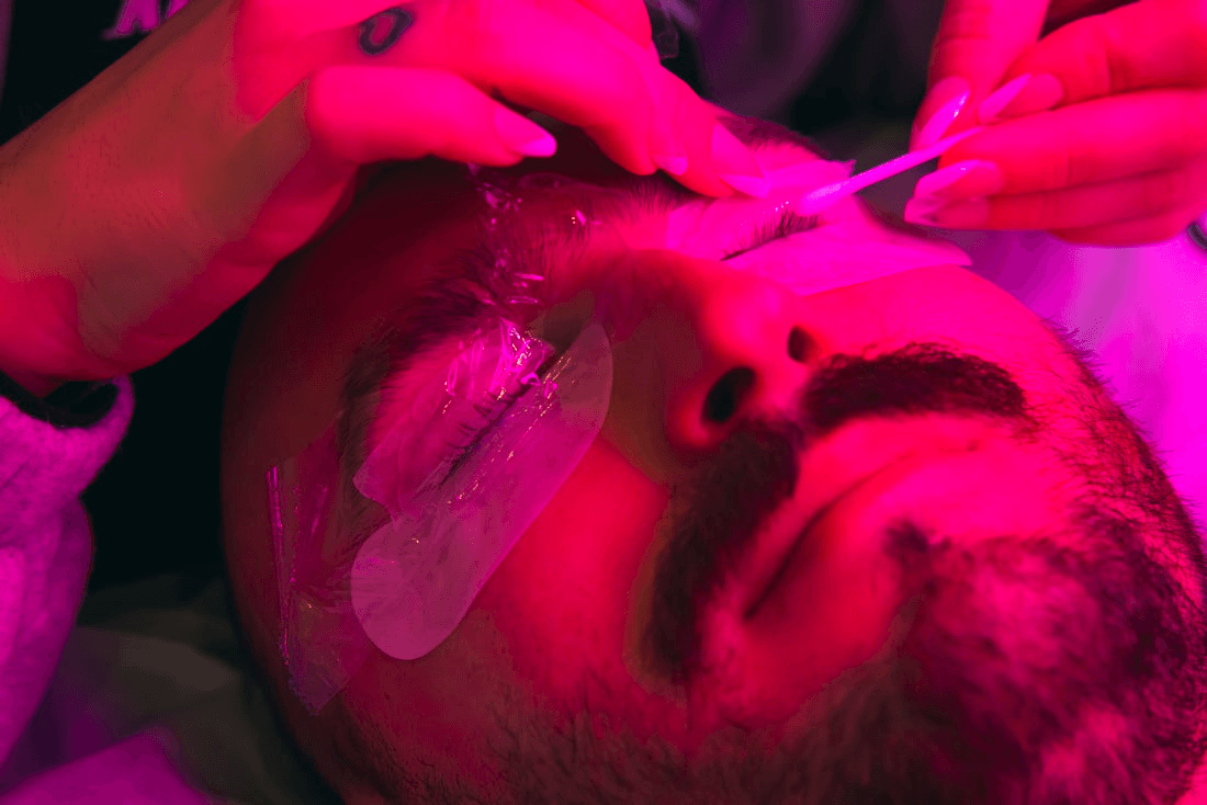 A person receives eyelash work from a stylist while bathed in pink light.