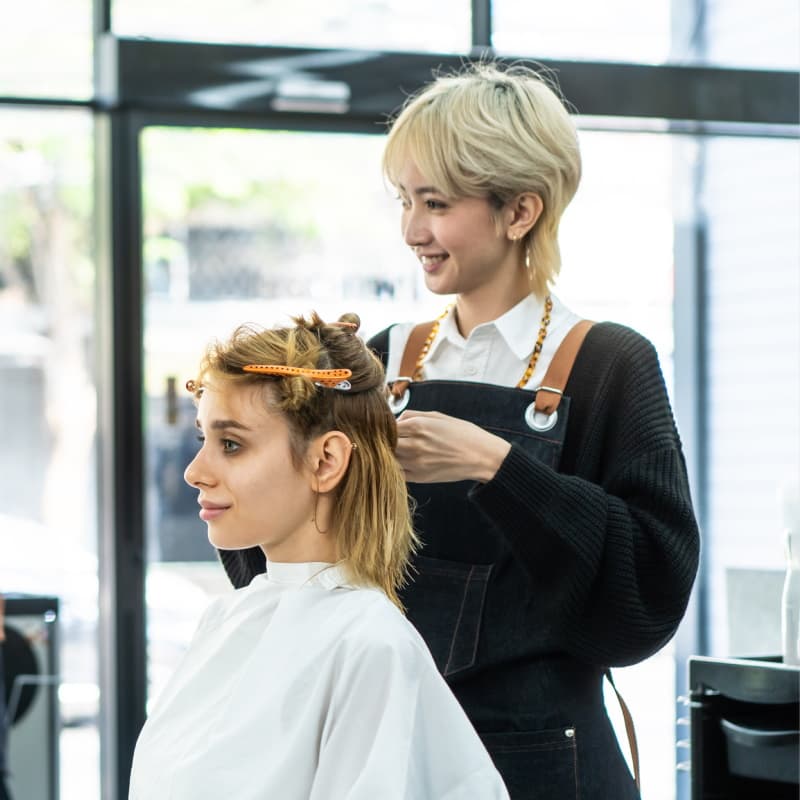 Two stylists cut hair in a well-lit salon, one in the foreground and one in the background.