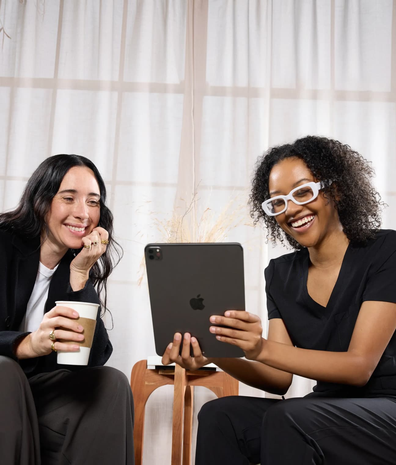 Two women sit together in a bright treatment room, smiling as they look at a tablet showing service options.