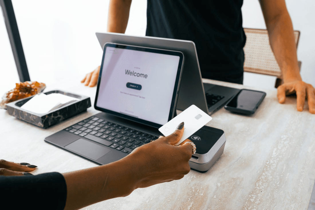 The front desk of a salon. Behind the counter, an employee stands with a laptop. The client faces a tablet and holds out a credit card.