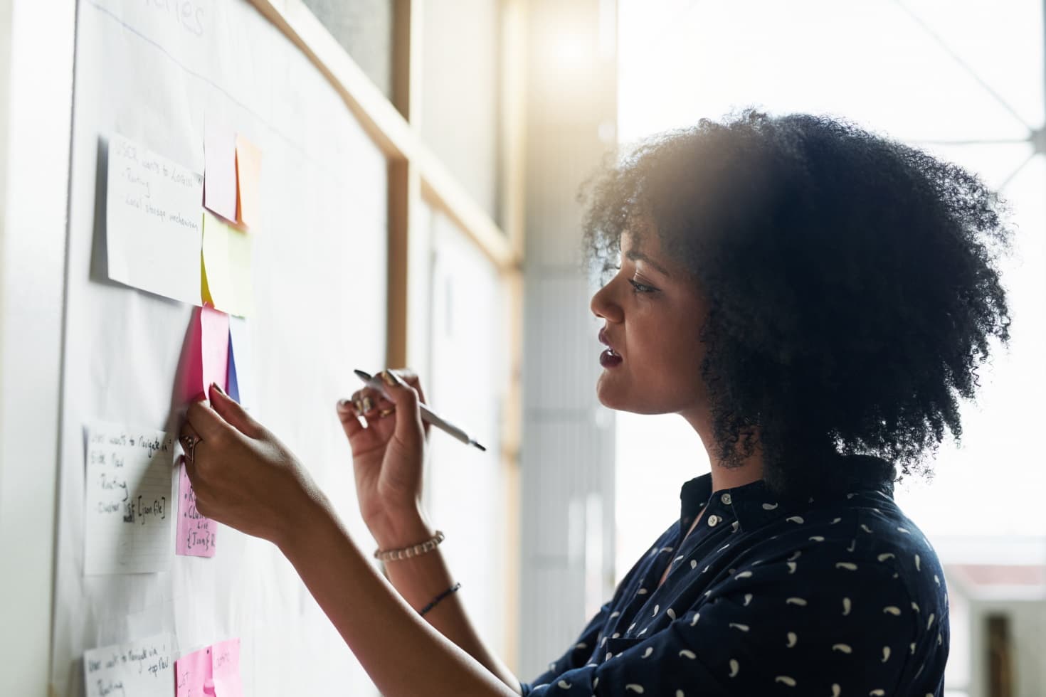 A young woman writes notes on colorful pieces of paper tacked to a bulletin board.
