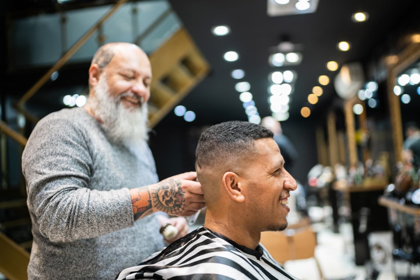 A barber with a bushy white beard grins while chatting with a client and trimming his close-cropped hair.